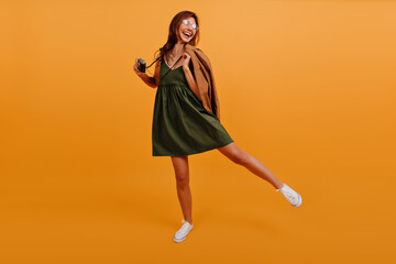 Canvas Print - Cheerful joyful girl in summer sundress is having fun and dancing on isolated background. Photo in studio model in full growth with camera in hand