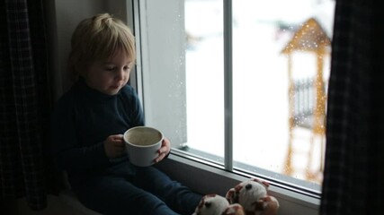 Poster - Toddler child, sitting on the window, watching the snow falling, reading little book