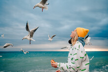 A Blonde Hipster Girl In A Yellow Hat Feeds Gulls On The Background Of The Sea