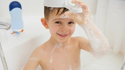 Wall Mural - Funny shot of little smiling boy having bath putting bucket with water on his head. Concept of child hygiene and health care at home. Child having fun and playing at home.