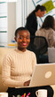© DC Studio - Portrait of african woman typing on laptop looking at camera smiling while diverse team working in background. Multiethnic coworkers talking about startup financial company in modern business office