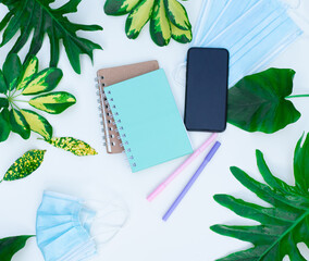 Notebook, phone, pen and green branches of tropical tree. Monstera leaves on white background with blue mask against coronavirus. business plan. flat bookmark, top view, flat lay