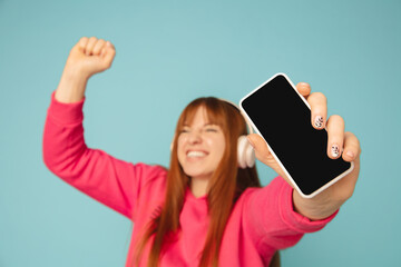 Wall Mural - Shows phone's screen. Caucasian woman's portrait on blue studio background with copyspace. Beautiful female model in pink sweatshirt. Concept of human emotions, facial expression, sales, ad, fashion.