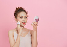 Young Attractive Asian Lady Applying Blusher On Her Face With Powder Puff Over Pink Background.