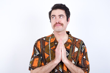 Charming cheerful Young caucasian man wearing generic pattern printed shirt against white wall making up plan in mind holding hands together, setting up an idea. Look askance