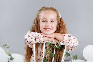 Cute little girl blonde smiling having fun in studio with white balloons and green. child in floral dress sitting on white chair and celebration easter. happy childhood