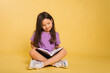 © Вероника Преображенс - beautiful little girl in a purple t-shirt reads a book while sitting on a yellow background. Cute child learns lessons.
