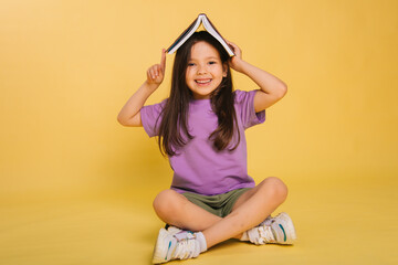 beautiful little girl holding a book on her head while sitting, playing while studying. Cute baby teaches lessons on a yellow background. Photo in studio