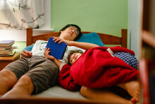 A Boy Cuddles Up To His Sleeping Father In Bed Reading Book At Bedtime
