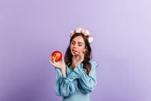 Girl In Great Mood Is Looking At Delicious Red Apple. Snapshot Of Woman In Blue Blouse On Isolated Background