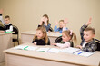 © nagaets - Group of school kids sitting in classroom and raising hands.