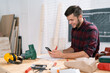 © ABCreative - Young bearded carpenter focused on planning woodworking project using cell phone in workshop