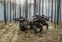 Squad Of Four Fully Equipped Soldiers In Camouflage On A Reconnaissance Military Mission, Aiming Rifles. They're Moving In Formation Through Dense Forest.