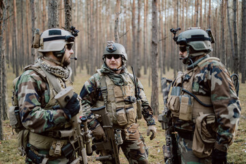 A group of middle-aged bearded soldiers in uniforms and tactical vests discusses the action plan and prepares for action at a temporary forest base. The commander using military tablet.