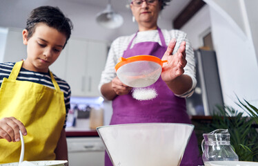 Caucasian mother teaching her son how to cook preparing the ingredients to make a cake. Child pouring flour to sift