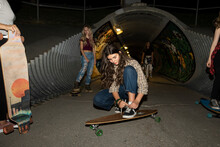 Teenage Girl Tying Shoe On Skateboard In Park With Friends At Night