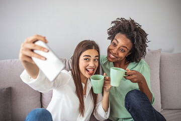 Wall Mural - Excited multicultural friends taking selfie and laughing. Indoor shot of two emotional caucasian and chinese girls. Two adult woman making selfies. Two female friends taking a selfie