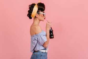 Sticker - Side view of pinup brunette girl drinking soda. Studio shot of stylish young woman in vintage outfit isolated on pink background.