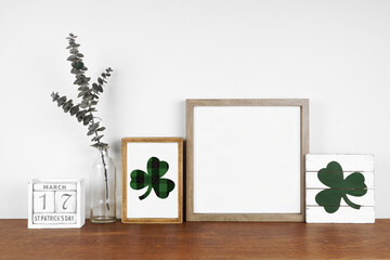 Mock up wood frame with St Patricks Day decor on a wood shelf. Shabby chic wood signs, calendar and green branches. Square frame against a white wall. Copy space.