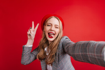 Wall Mural - Carefree girl in elegant jacket taking selfie. Funny french woman showing peace sign on red background.