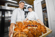 © salomonus_ - Bakers man and girl with a full box of hot pastries in their hands ha the background of an industrial oven in a bakery.