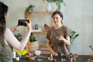 Wall Mural - Young woman with small bowl containing essence oil looking at smartphone camera