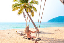 Blonde Woman With Long Hair Swinging On A Swing Suspended From A Palm Tree Near The Sea In Thailand, Rear View, Real People. Happy Vacation In Asia.