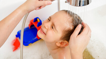 Little boy closing his eyes while mother washing his head with shampoo under shower in bath. Concept of child hygiene and health care at home.