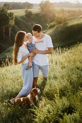 Young beautiful family with a little daughter and a dog hug, kiss and walk in nature at sunset.