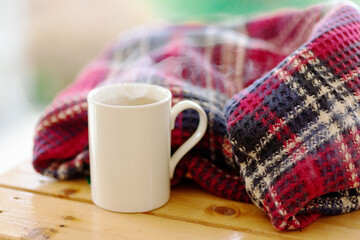 Autumn, winter cozy composition. Plaid, steaming white color cup of hot tea on wood table.concept of coziness and interior. Front view, copy space,mock up.