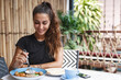 © Liubov Levytska - Food and tropical vacation concept. Beautiful tanned woman eating at hotel cafe, sit outdoors on terrace and trying Bali breakfast, drinking morning coffee and smiling