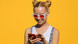 Portrait of happy teenager blonde girl in wireless headphones in sunglasses listens to music on smartphone, smiles, dances, shaking her head rhythmically against  yellow background summer