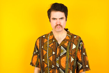 Wall Mural - young Caucasian man wearing printed shirt against yellow wall frowning his eyebrows being displeased with something.