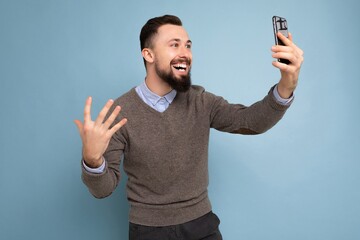 Side-profile Photo of positive handsome young brunette unshaven man with beard wearing casual grey sweater and blue shirt isolated on pink background wall holding smartphone having video call looking