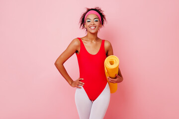 Fascinating black woman posing in red aerobics form. Studio shot of positive african girl with yellow mat.