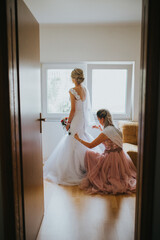 Poster - Vertical shot of a bride and bridesmaid with pink dress helping the bride to wear her wedding dress