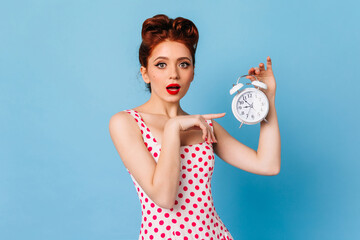 Surprised woman with bright makeup showing time. Studio shot of shocked pinup girl holding clock on blue background.