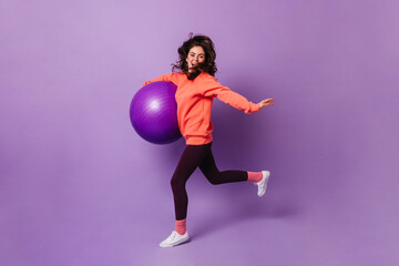 Canvas Print - Nice girl in pink socks, orange sweatshirt and dark leggings runs on purple background with fitball