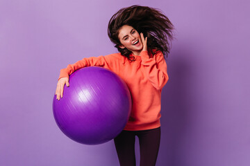 Canvas Print - Smiling lady in bright sweatshirt and dark leggings jumps on purple background, holding fitball