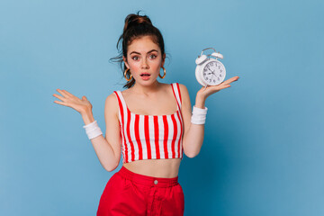 Canvas Print - Girl with ponytail and nude make-up surprised looking at camera on blue background. Woman in clothes in style of 80s posing with alarm clock