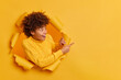 Positive beautiful curly haired woman with glad expression points away on copy space advertises something breaks through paper hole wears casual yellow sweater. People and promotion concept.