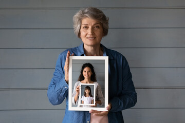 Head shot portrait smiling mature woman holding photo frame of grownup daughter and little granddaughter in row, standing on grey wooden wall background isolated, three generations of women concept