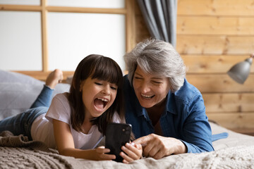 Wall Mural - Close up overjoyed little girl with mature grandmother using phone together, lying on cozy bed at home, spending leisure time with gadget, having fun, browsing apps, chatting online, watching video