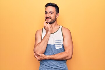 Wall Mural - Young handsome man with beard wearing sleeveless t-shirt standing over yellow background smiling looking confident at the camera with crossed arms and hand on chin. Thinking positive.