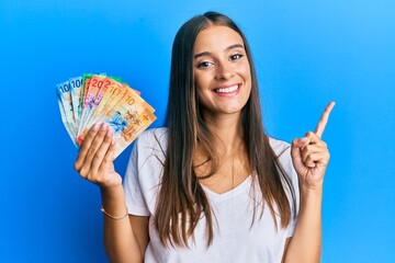 Wall Mural - Young hispanic woman holding swiss franc banknotes smiling happy pointing with hand and finger to the side