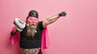 Surprised bearded man keeps mouth widely opened stretches tattooed arm holds tool for cleaning pretends to be superhero ready for actions isolated over pink background with copy space aside.