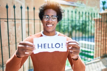 Wall Mural - Young handsome african american man smiling happy. Standing with smile on face holding hello message paper at town street.