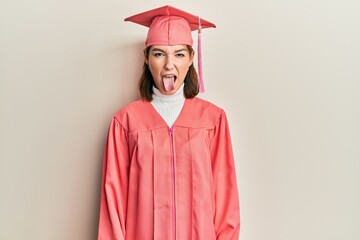 Young caucasian woman wearing graduation cap and ceremony robe sticking tongue out happy with funny expression. emotion concept.