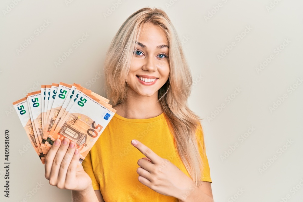 Beautiful caucasian blonde girl holding bunch of 50 euro banknotes smiling happy pointing with hand and finger - obrazy, fototapety, plakaty 