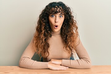 Young hispanic girl wearing casual clothes sitting on the table scared and amazed with open mouth for surprise, disbelief face